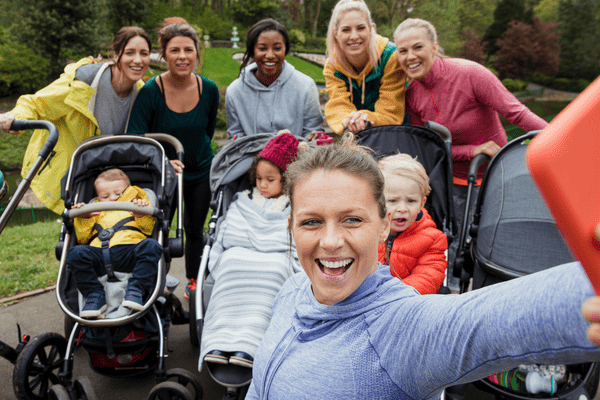 A woman takes a selfie-style photo of a group of mothers with babies in prams
